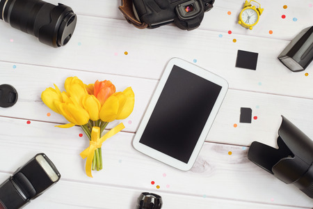 Workplace photographer: camera, lens, flash, memory card, alarm clock, tablet and a bouquet of tulips on a wooden table. Photographer Day concept.の写真素材