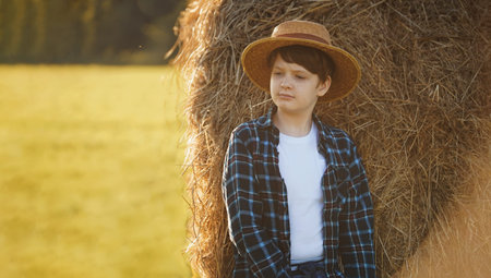 Teenage boy in straw hat standing near hay bale in a field, harvest concept.