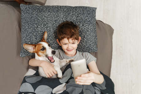 Small happy boy lies on a bed with jack russell terrier dog and takes a selfie.の写真素材