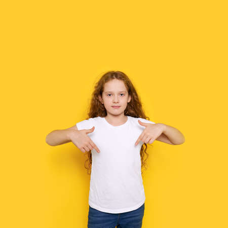 Curly child pointing his fingers on a blank t-shirt, on yellow background with a place for your advertising.の写真素材