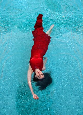 beautiful young woman in red evening dress elegant floating weightlessly in the pool in turquoise waterの写真素材