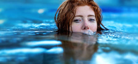 Portrait of young sexy playful woman with red hair, redhead, makes funny bubbles into water with mouth, swimming in spa pool, head half submerged under water, copy spaceの写真素材