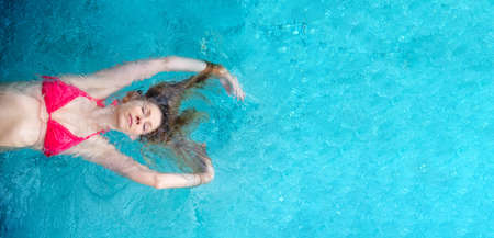 young sexy blonde woman in a pink bikini, swimming in the turquoise spa pool, long hair around her arms, copy spaceの写真素材