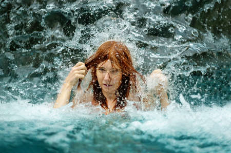 beautiful sexy redhead woman in bikini under the splashing water shower of the waterfall in the spa wellness pool strokes her hair with her hands and enjoys the falling water, copy spaceの写真素材
