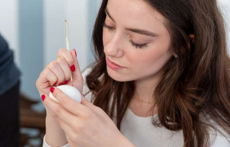portrait of beautiful, young, sexy, brunette woman paint a white Easter egg according to Sorbian tradition with a needle head quill and melted candle wax, copy spaceの写真素材