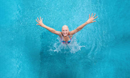 Top view beautiful young sexy blonde playful woman, in colorful bikini, in the turquoise spa wellness pool, raises her arms, jumping happy, laughing out of water, splashing the pool water, copy spaceの写真素材