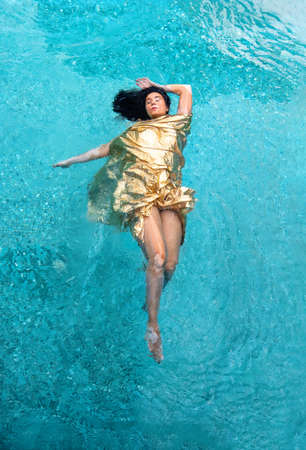 Top view of a beautiful young sexy mermaid woman with vitiligo pigment spot disease in golden dress, evening dress cape, floating weightless elegant swimming in turquoise spa pool water, copy spaceの写真素材