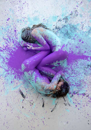Two artistically abstract painted young sexy women, girlfriends in pink, purple, turquoise and white color lying like a Yin Yang symbol on the floor in the artists studio, copy space.の写真素材