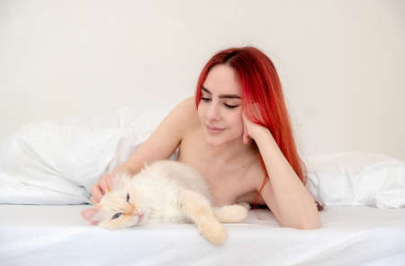 Portrait of an attractive, content, young, sexy red-haired woman lying relaxed in bed, enjoying and snuggling with fresh, soft, white bedding and cat in the bedroom.の写真素材