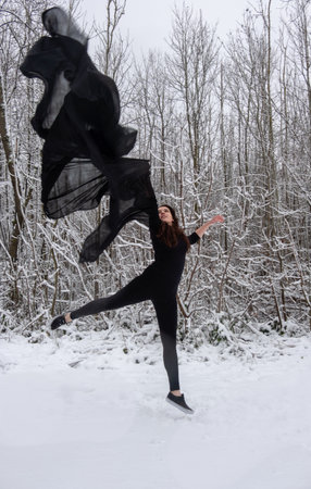 Young woman in black ballet suit jumping with joy in the snowy forest with a black cloth in the airの写真素材