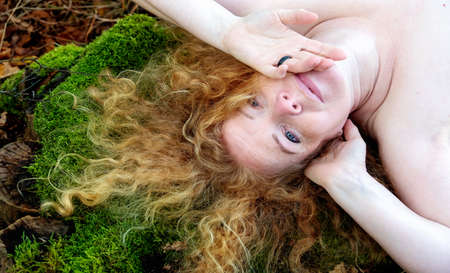top view portrait of a beautiful sexy lovely mature redhead woman with long red curly hair lying on green moss trunk in the forestの写真素材