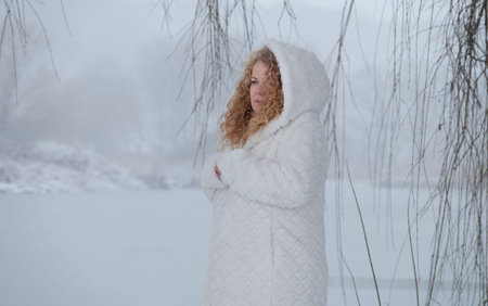 redhead mature woman in her fifties with red curly hair and white hooded coat freezes in the snowy...