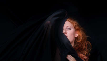 Portrait of a beautiful seductive sensual, young sexy redhead, a beautiful ginger woman with long curly red hair with black cloth over head and folded hands, copy spaceの写真素材