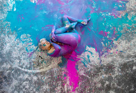 Young naked woman in turquoise and magenta paint color, painted, lies decorative, elegantly curled up in fetus pose on the colorful studio floor. Creative, abstract expressive body art and painting.の写真素材