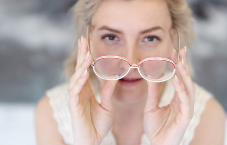 Portrait of a young woman with glasses and blond hair holding the glasses in front of her face with...