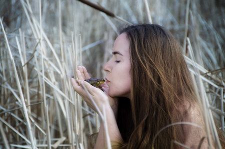 Young cute woman in reed kisses a frog, enchanted prince like in fairy tale frog prince, copy space