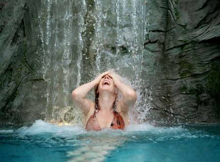 beautiful young sexy redhead woman in bikini under the splashing water of the waterfall in the spa wellness pool raises her arms, laughing and having fun, copy spaceの写真素材