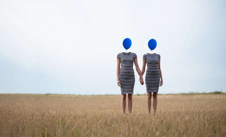 surreal image of two women standing hand in hand on a field, their heads are replaced by blue balloons, copy spaceの写真素材