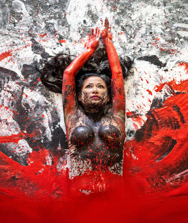 nude artistic asian woman in red and black paint, painted, lying on the patterned ground, the lower body emerges, arises from red color. Creative, abstract body art, colorful painting, body painting.の写真素材