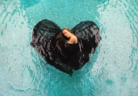 Top view from above to a beautiful sexy young woman floating in black cloth dress wrapped in shape of valentine heart in turquoise blue water of the swimming pool, copy spaceの写真素材