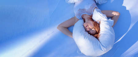 wide banner view from above to an attractive, young, sexy redhead woman, enjoys the fresh soft white bed, in nightwear, underwear, in the bright morning sun, blue shadows, copy space.の写真素材