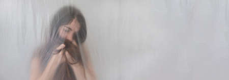 blurred, fuzzy wide panoramic banner portrait of depressed sad young woman with long hair puts head in hands, behind transparent plastic foil film, copy spaceの写真素材