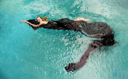 Top view to a beautiful sexy young woman floating like a mermaid in black cloth dress wrapped in green water of the swimming pool, elegant erotic dream, copy spaceの写真素材