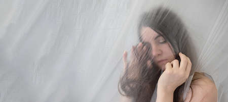 blurred, fuzzy image of sensual romantic, sad young woman with long hair puts head in hands, behind transparent plastic foil film, copy spaceの写真素材