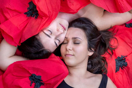 two beautiful young sexy brunette women girlfriend, with closed eyes lie together with sensual touch, lying covered with fashion red paper poppies, copy spaceの写真素材