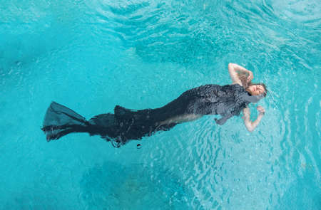 Top view to a beautiful sexy young woman floating like a mermaid fish, wrapped in black cloth dress in turquoise blue water of the swimming pool, elegant erotic dream, water ballet, copy spaceの写真素材