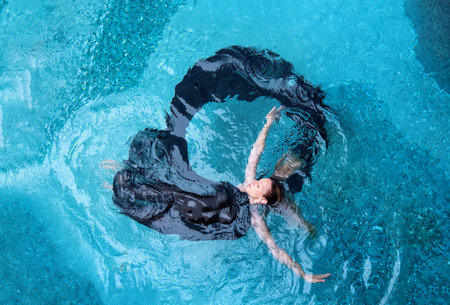 top view from above to a beautiful sexy young woman floating, wrapped in black cloth dress in the form, shape of a ring, circle in turquoise blue water of the swimming pool, copy spaceの写真素材