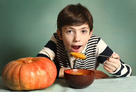 preteen handsome boy with big orange pumpkin eat mash cream soup close up portrait on blue wall backgroundの写真素材