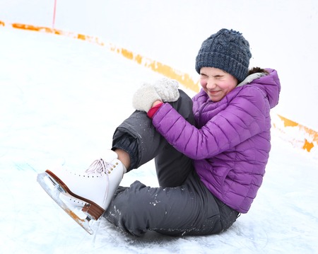 teen skater girl in down jacket and knitted blue hat fall down on skating rink hit her leg and grimace with painの写真素材