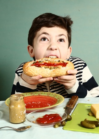 preteen handsome boy cook by himself big hotdog from french baguette sausage cucumber cheese ketchup and mustard and make big biteの写真素材