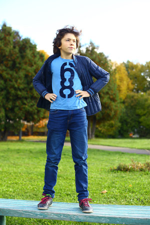 preteen hansome boy in blue jeans and jacket on the fall background in the city park stand on the benchの写真素材
