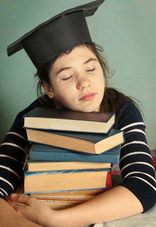 intelligent teen girl in graduation cap wiht book pile sleeping tired of studyingの写真素材