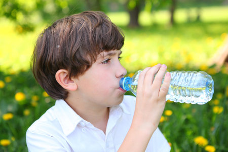 preteen handsome boy drink water. Boy drinking water from the bottle on the outdoors summer park green background.の写真素材