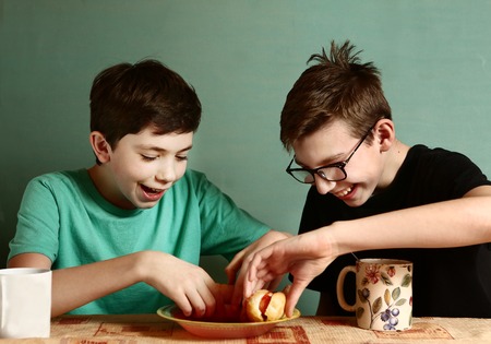 teenager school boys cooking eat hot dog happy smiling laughing closeup portrait on blue wall backgroundの写真素材
