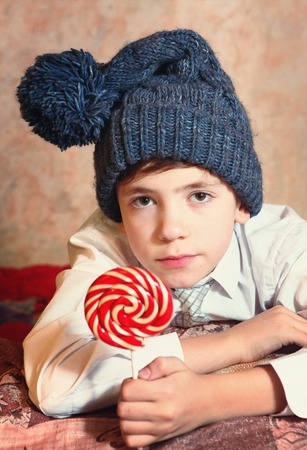cute preteen boy in blue knitted hat with red and white round candy on stickの写真素材