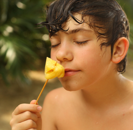 teenager boy eat pineapple piece on stick close up outdoor photo on tropical palm beach backgroundの写真素材