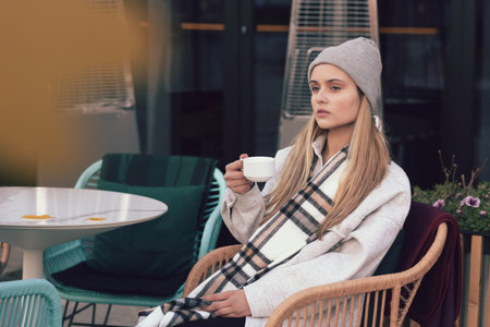 blond lgirl in knitted hat and scarf with cup of tea close up portrait in cafeの写真素材