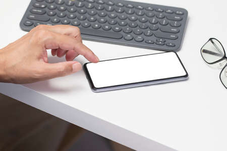 Hand touching smartphone with blank screen, with office desk environmentの写真素材