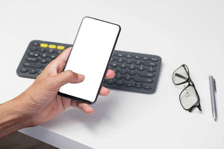 Hand touching smartphone with blank screen, with office desk environmentの写真素材