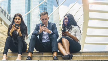 group of three people multi nationality Business workers man and woman use mobile phone and sit on the stair talking about they work in the morning with the city space backgroundの写真素材