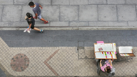 people walk on the pedestrian concrete pavement with the disabled lady on the wheel chair sells the lottery on panel stand.の写真素材