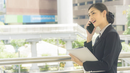 Asian business woman uses and talks on mobile phone with paper work on her hand at outdoor の写真素材
