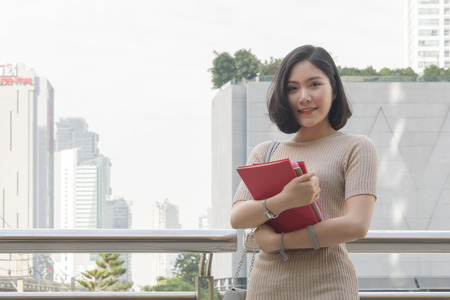 portrait of asian teen girl holds red book stands and dresses up in street fashion cloth at outdoor public space with modern building backgroundの写真素材