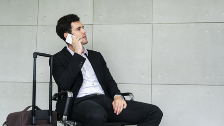 business man is sitting on chair waiting for business trip travel with suitcase and calling smartphoneの写真素材
