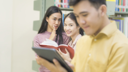asian teenager man reads a tablet with girls talk at the book shelf in the background. (focus at girls)の写真素材