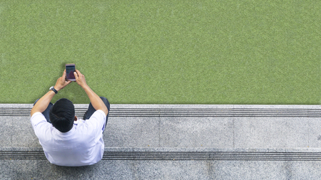 the top aerial view of the man in white shirt uses mobile phone and sits on the pedestrian concrete walk way at glass green landscape.の写真素材